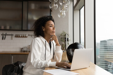 Close up thoughtful African American businesswoman looking out window, touching chin, sitting at wooden table with laptop and motorcycle helmet, pondering project strategy, planning, visualizing