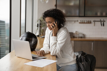 Confident African American businesswoman making phone call, using laptop, searching information, consulting client, negotiating with business partner, sitting at table with motorcycle helmet