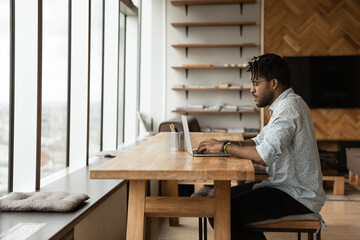 Side view African American man using laptop, sitting at wooden table in modern apartment, confident...