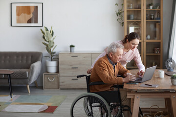 Side view portrait of senior man in wheelchair using laptop at retirement home with nurse assisting him, copy space