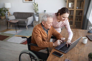 Portrait of senior man in wheelchair using laptop at retirement home with nurse assisting him, copy...