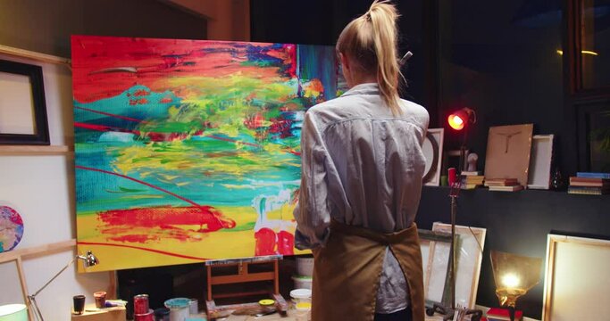 Back view of young Caucasian blonde talented female painter looking at finished oil painting standing in art studio alone Art workshop small business, contemporary abstract artwork, creativity concept