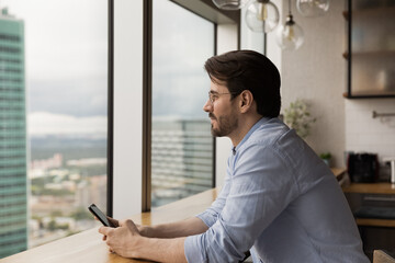 Fototapeta premium Side view close up dreamy man wearing glasses distracted from phone, looking out window, waiting for call, pensive businessman visualizing new opportunities, dreaming about good future, planning