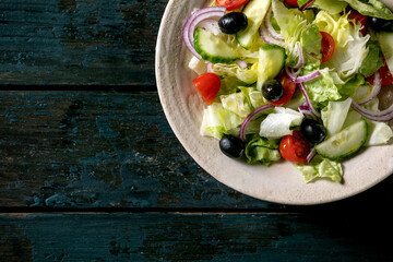Classic vegetable salad with tomatoes, cucumber, onion, salad leaves and black olives in white ceramic plate. Dark wooden background. Flat lay, copy space