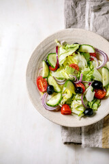 Classic vegetable salad with tomatoes, cucumber, onion, salad leaves and black olives in white ceramic plate on cloth napkin. White marble background. Flat lay, copy space