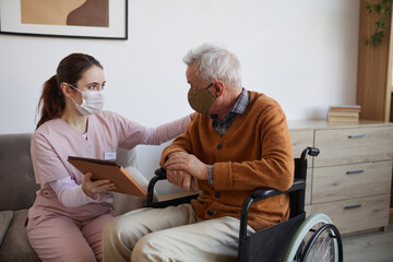 Portrait of young nurse assisting senior man in wheelchair using digital tablet at retirement home, both wearing masks, copy space