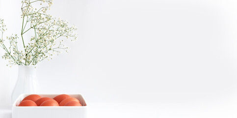 Easter eggs in a white square dish, on a white table, and a vase with delicate white flowers. Banner, space for text