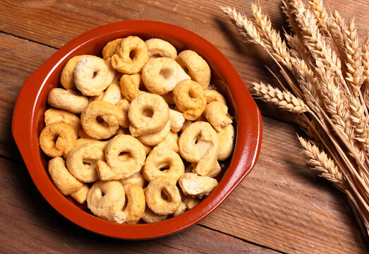 Ciotola marrone su sfondo di legno con taralli pugliesi e a fianco spighe di grano.