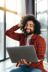 African American man in a red plaid shirt sits in an apartment near a brick wall working at a laptop