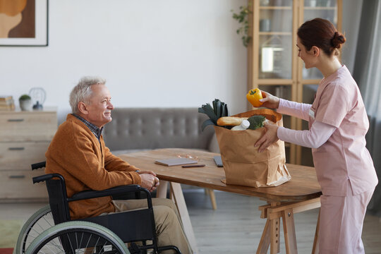 Side view portrait of smiling senior man in wheelchair looking at female nurse bringing groceries, assistance and food delivery concept