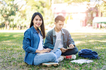 asian student couple studying outside on the grass, sitting down on the school campus using laptop computer, books, pencil and note book, studying hard under the trees nature in the evening sunlight