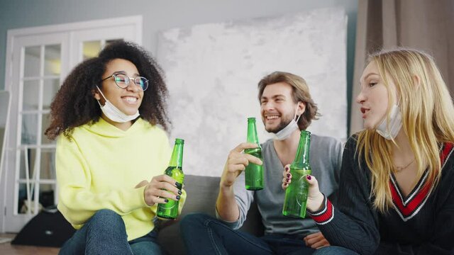 Mixed race group of student friends drink beer indoor with masks pulled down around chin during the coronavirus pandemic