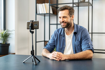 Smiling mature handsome man blogger, stylish, sitting in front of cellphone on tripod stabilizer...