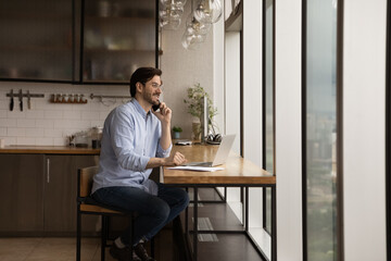Full length smiling businessman wearing glasses talking on smartphone, looking out window, sitting at table with laptop at home, manager consulting customer by phone call, young man chatting