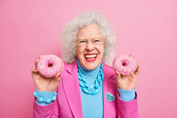 Charming happy mature woman smiles broadly shows white perfect teeth holds two glazed doughnuts...