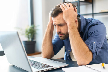 Terrified young adult businessman terrified looking at computer screen, frowning, holding the head...