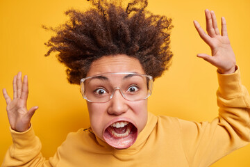 Close up portrait of emotional curly haired woman stares at camera has very huge mouth raises arms wears optical big glasses has hair raised isolated over yellow background. Madness emotions