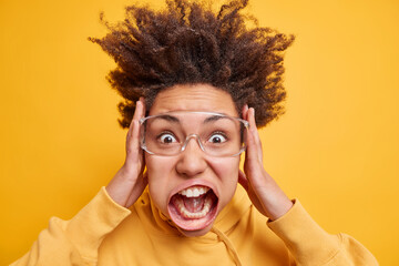 Portrait of emotional shocked curly haired woman grabs face and shouts loudly keeps mouth widely...