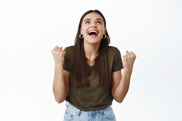 Happy and relieved young woman scream of joy and happiness, winning prize, triumphing, achieve goal or prize, raising hands up and looking at top, white background