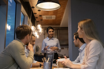 Confident Indian businesswoman mentor leading corporate briefing in evening, speaking, explaining project strategy, diverse colleagues involved in brainstorm at meeting, business partners negotiation