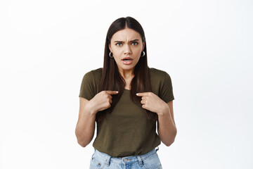 Shocked and offended young woman pointing at herself with displeased face, being insulted, feeling...
