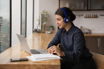 Side view Indian woman wearing headphones looking at laptop screen, student or intern watching webinar, training, listening to lecture, taking notes, sitting at wooden table, studying online