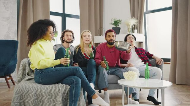 Multiethnical young and cheerful men and women watching comedy movie on TV together in the loft styled living room with beer and snacks and laughing laud. Favorite TV series