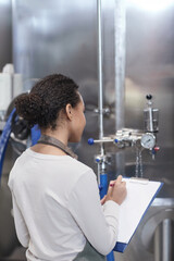 Vertical back view portrait of young African-American woman holding clipboard while inspecting production quality at industrial food factory
