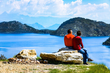 Fototapeta na wymiar A couple sitting on a big rock and enjoying the view of the blue waters of the Mediterranean sea laying before them