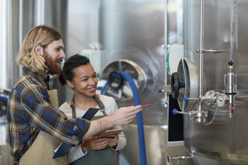 Side view portrait of two smiling young workers inspecting production at modern craft brewery, copy...