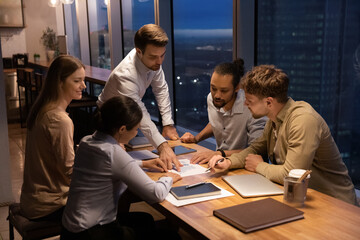 Diverse employees working with financial project statistics at late meeting in office, confident businessman executive pointing at document with diagrams and charts, multiracial team brainstorming