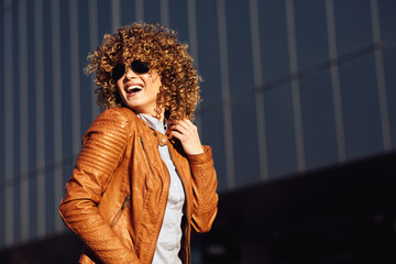 Portrait of a young happy woman in a brown leather jacket with blond afro curly hair and wears sunglasses on the street © djile