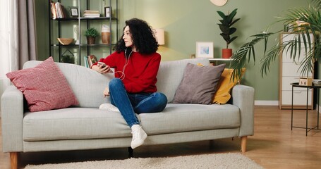 Caucasian pretty young happy brunette woman sitting on sofa in cozy living room listening to music in earphones on smartphone. Young female resting at home relaxing enjoying favorite songs