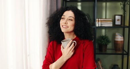 Fototapeta na wymiar Close up portrait of cheerful happy dreamy Caucasian beautiful young woman standing near window in room looking away with smile on face holding cup of coffee or tea dreaming of something, home concept