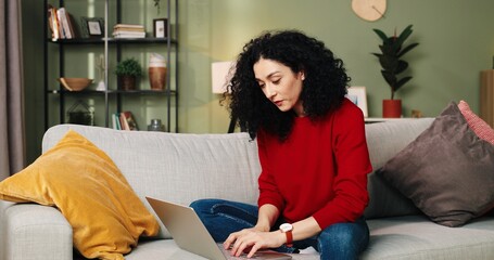 Caucasian beautiful happy young female typing browsing online on laptop, using social media sitting on couch at home, student studying on computer in living room using digital gadget