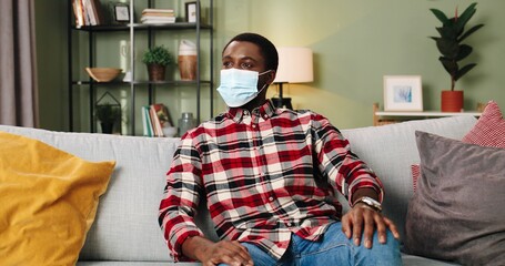 Portrait of African American young handsome man in medical mask looking at camera while sitting alone in living room in apartment in quarantine during coronavirus. Home concept, leisure time