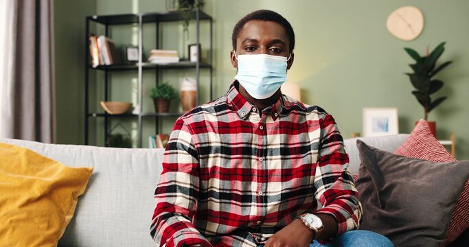 Close Up Portrait Of Young Happy African American Man Sitting At Home On Sofa Taking Off Protective Mask And Looking At Camera Smiling.