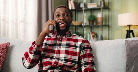 Portrait of Cheerful young African American handsome man holding smartphone talking on call while sitting on couch in living room. Happy guy speaking calling on cellphone resting in cozy apartment