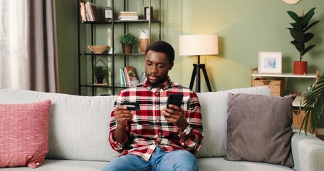 Obraz premium Close up. Camera approaching African American excited happy young handsome male sitting on sofa at home and buying online e-commerce shopping on internet using smartphone. Buyer concept