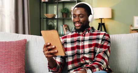 Young African American man in white headphones sits on sofa at home and chats via video with...