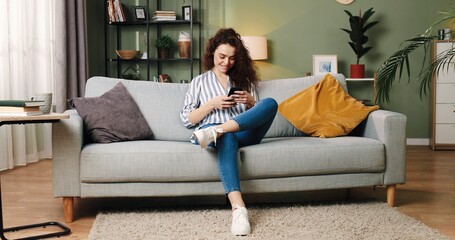 Caucasian pretty young cheerful smiling female sitting on sofa and texting on smartphone resting in room at home browsing online searching internet, social network user, mobile phone application