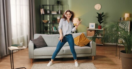 Happy joyful young Caucasian beautiful woman in positive mood dancing moving rhythmically and jumping while listening to music song in headphones on smartphone, having fun in room, leisure concept