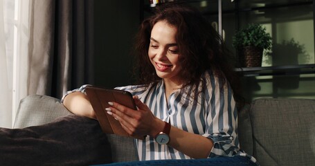 Close up of happy Caucasian cheerful young female smiling in good mood sitting in apartment alone typing on tablet device surfing internet with dreamy face, positive emotions, Leisure concept