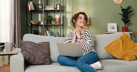 Caucasian beautiful young smiling joyful female sitting in room on sofa and typing searching...