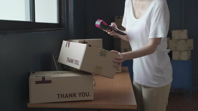 Asian Female seller online store worker holding scanner scanning parcel bar code packing e commerce post shipping box preparing online retail shop order in drop shipping delivery service warehouse.