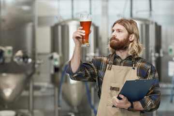 Waist up portrait of bearded brewmaster holding beer glass while inspecting quality of production...
