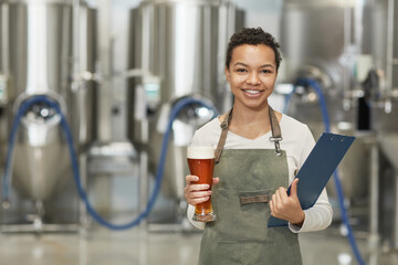 Waist up portrait of smiling African-American woman holding beer glass and looking at camera while...