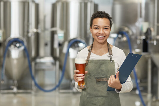 Waist up portrait of smiling African-American woman holding beer glass and looking at camera while standing in workshop at brewing factory, copy space