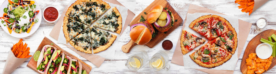 Healthy plant based fast food table scene. Overhead view on a white wood banner background. Cauliflower crust pizzas, bean burgers, mushroom tacos and vegetarian sides.