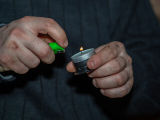 A man with dirty, frozen, soot-stained hands lights the wick of a decorative candle with a lighter. The concept of frostbite of the limbs, overcoming, fighting for life during the war.